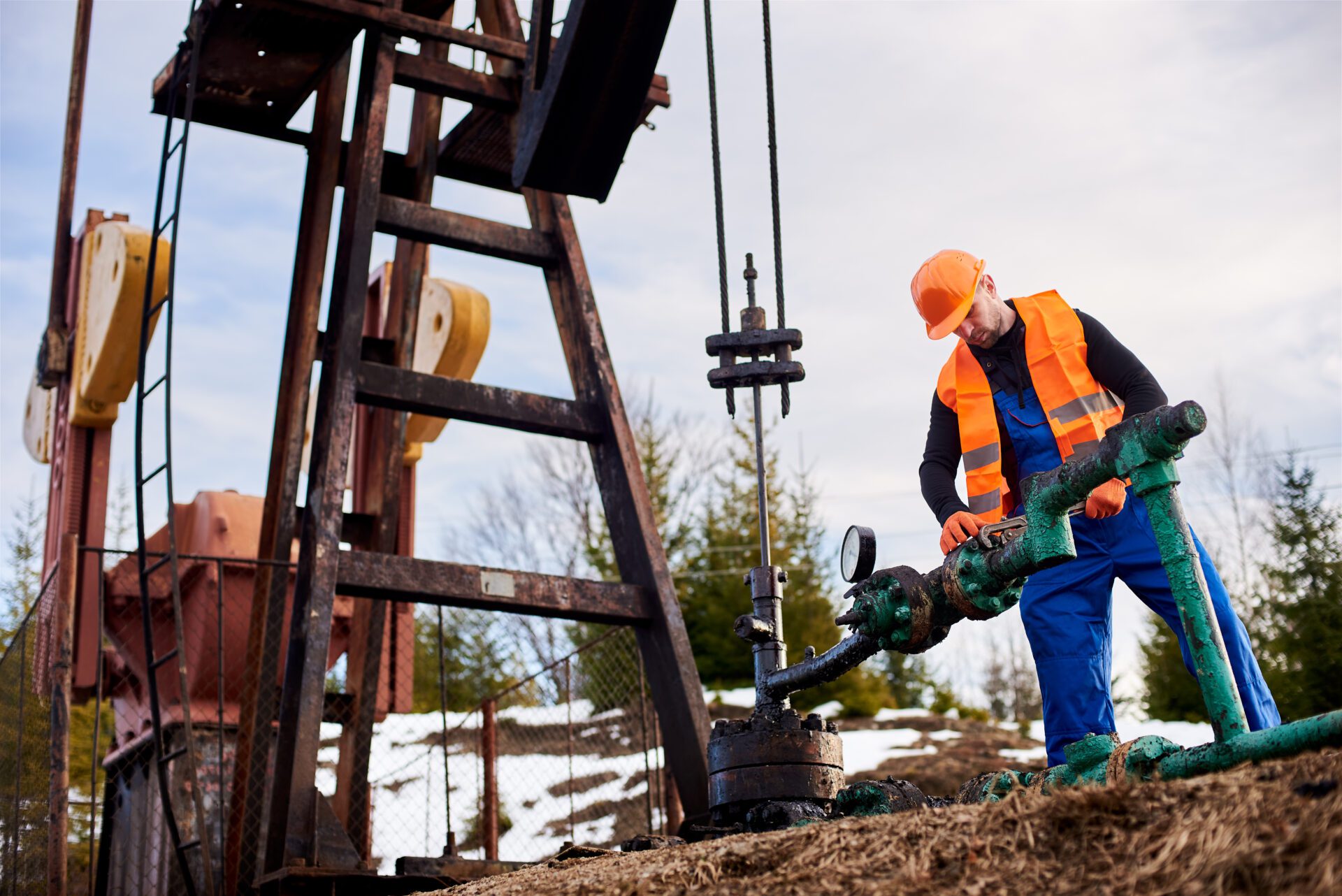 Well worker in blue overalls, orange vest and helmet using industrial wrench while repairing pipe of oil well pump jack, tightening bolt on pipe